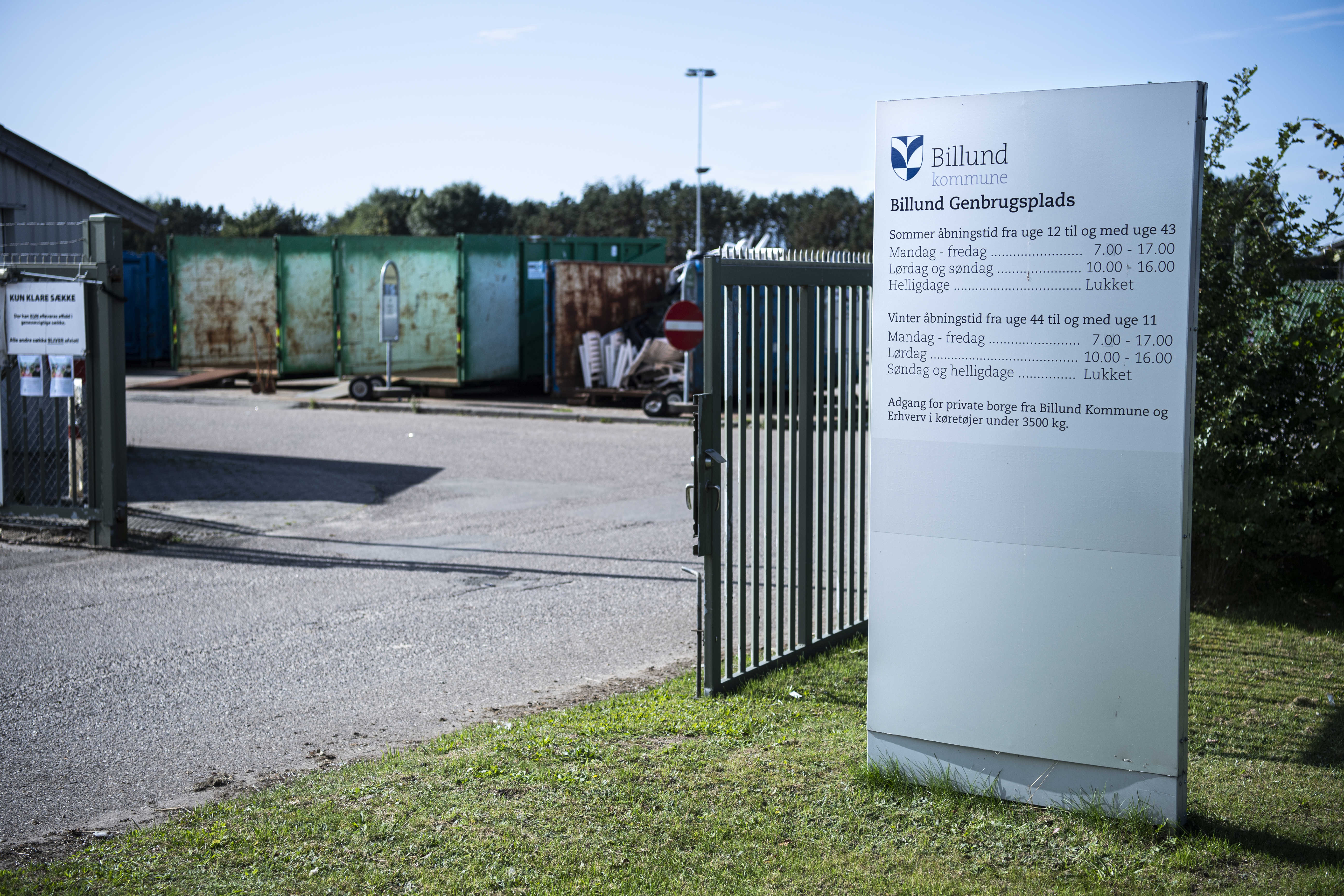 Indgang til Billund Genbrugsplads med en stor informationsstander, der viser åbningstiderne. En åben port leder ind til pladsen, hvor flere store containere til affaldssortering er synlige i baggrunden. Skilte og afspærringer markerer adgangsforholdene.