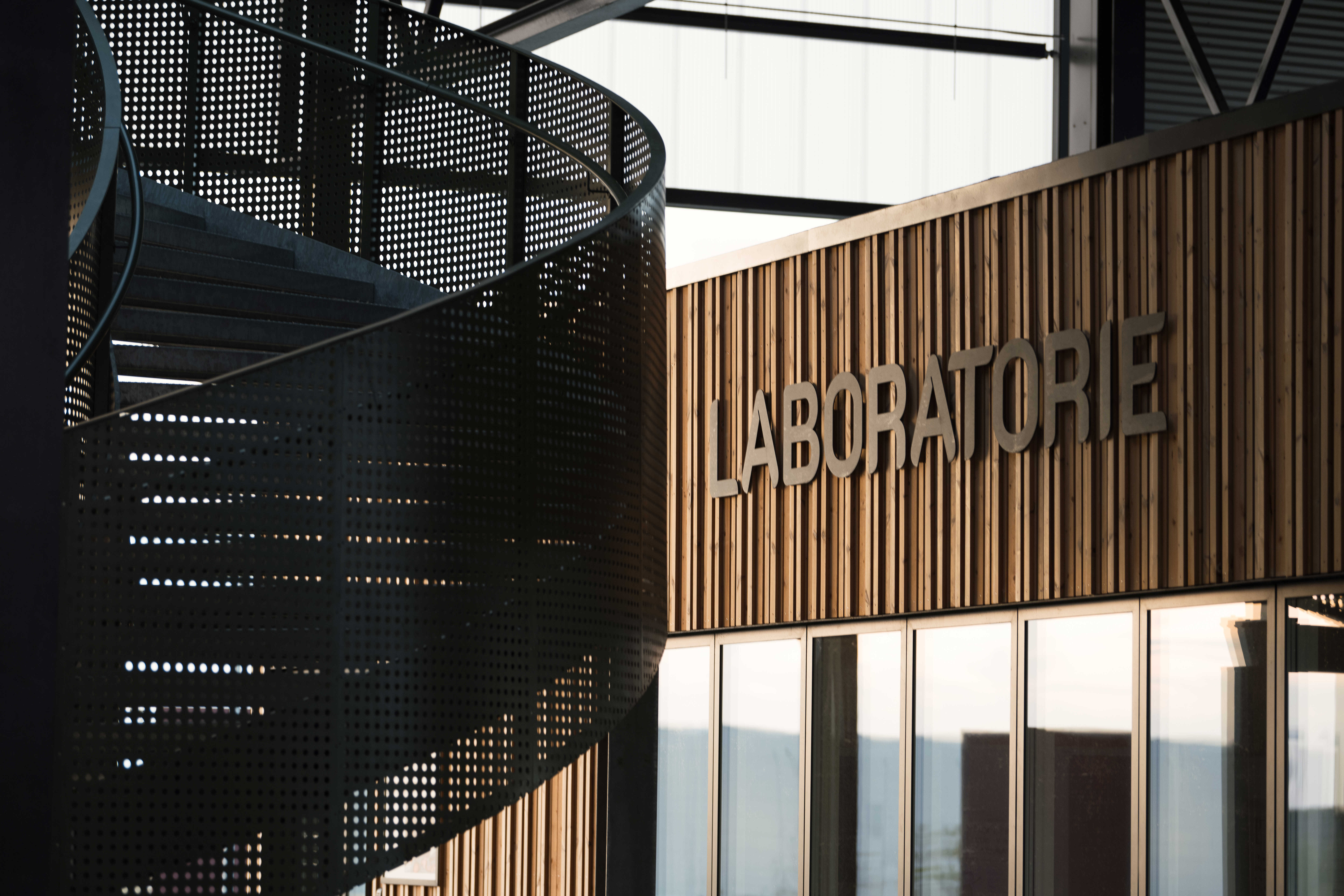 Nærbillede af facaden til Laboratoriet på RessourceCenter Vejle (RCV). Facaden er lavet af trælameller, og ordet "LABORATORIE" er monteret i store bogstaver. Til venstre ses en moderne vindeltrappe i perforeret metal, der tilføjer et industrielt præg til designet.