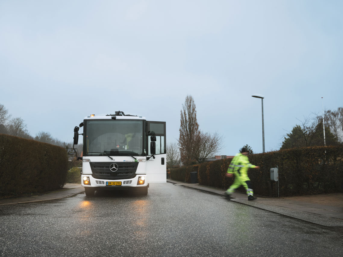 Skraldebil set forfra på en regnfuld dag, mens en skraldemand krydser vejen