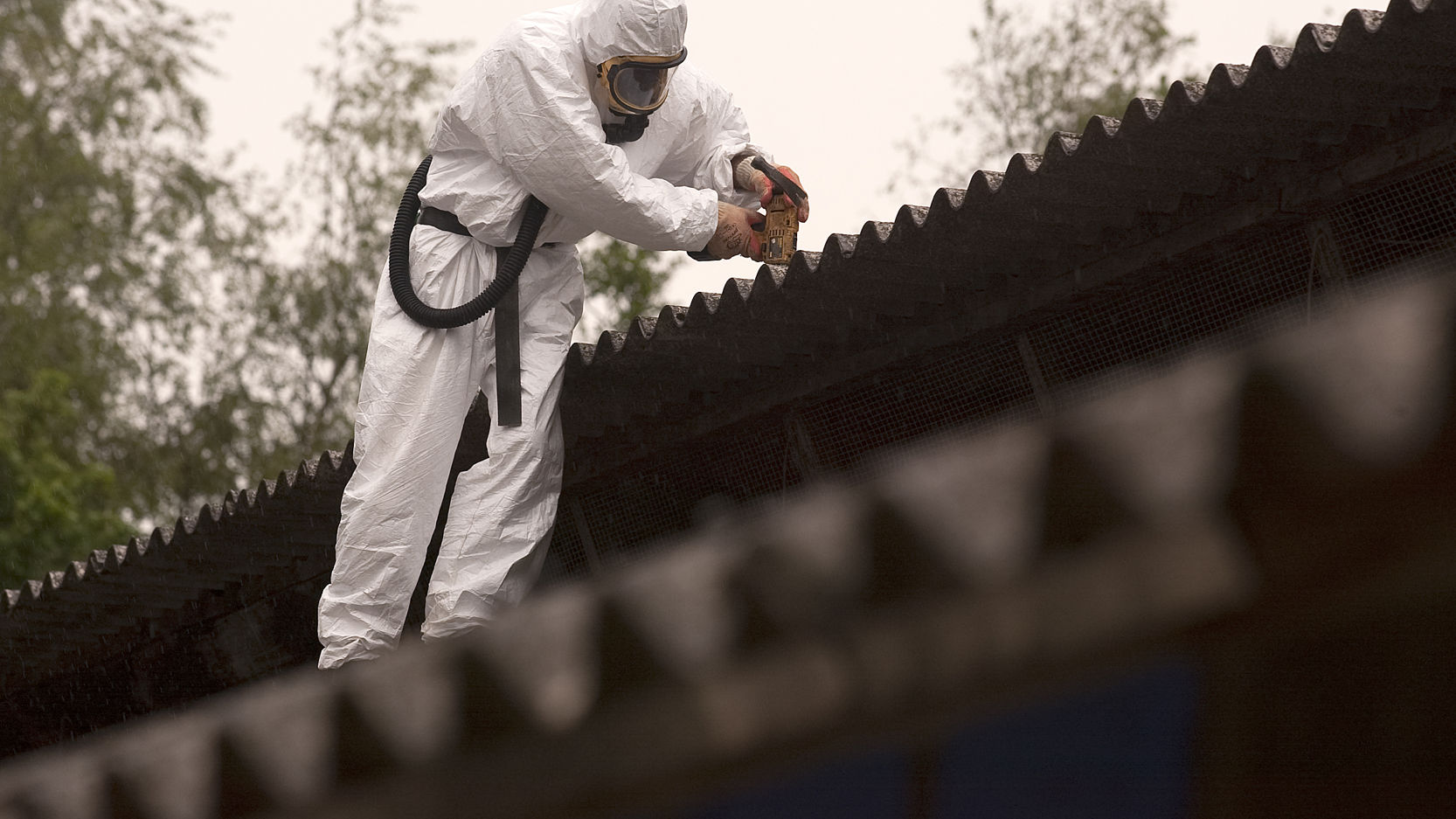 Person iført sikkerhedsudstyr og åndedrætsværn i færd med at renovere et asbest-tag.