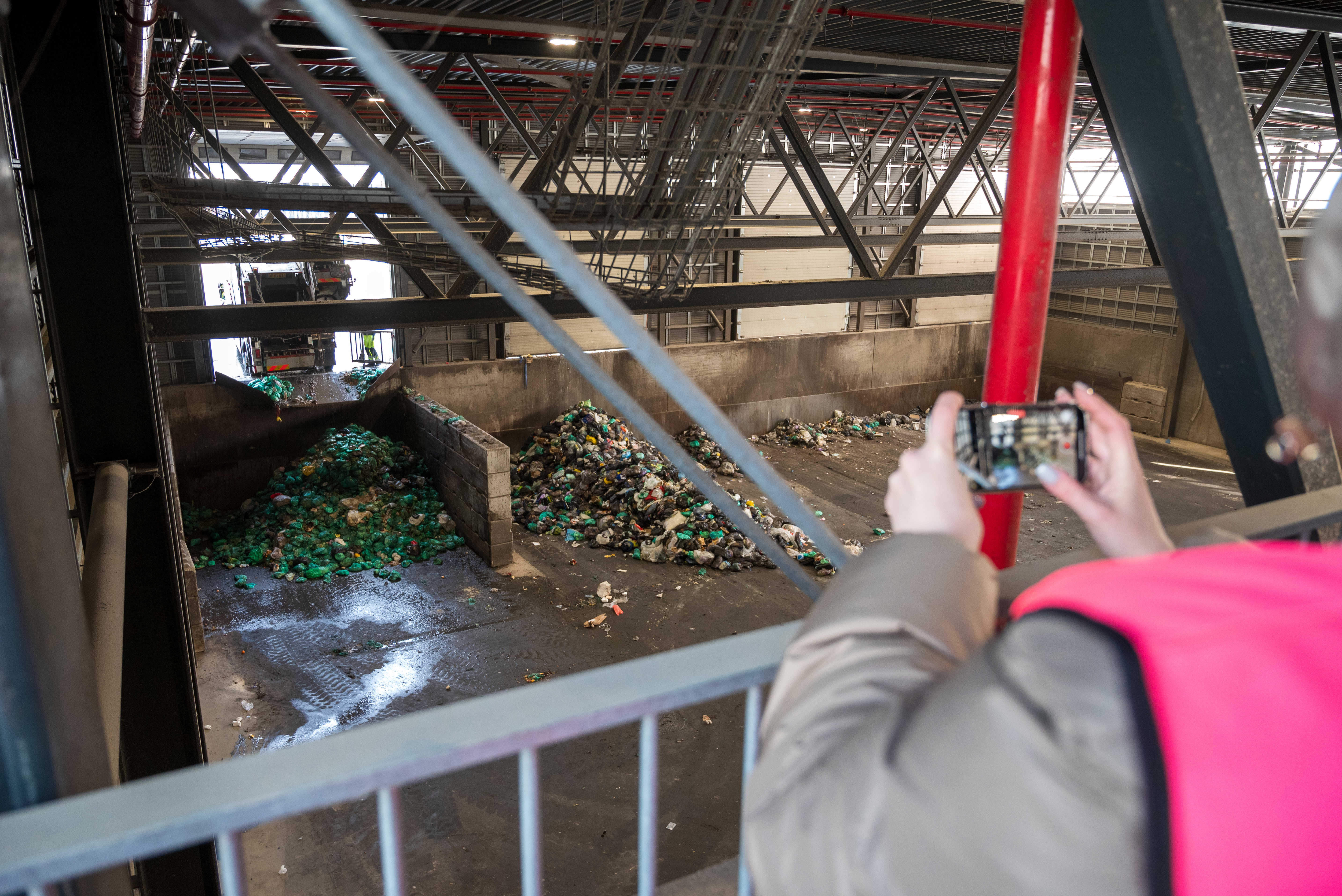 En person iført pink sikkerhedsvest tager fotos med en iPhone fra en af platformene på Læringsbroen med udsigt ned til en lastehal, hvor en renovationsbil er i gang med at læsse madaffald af.