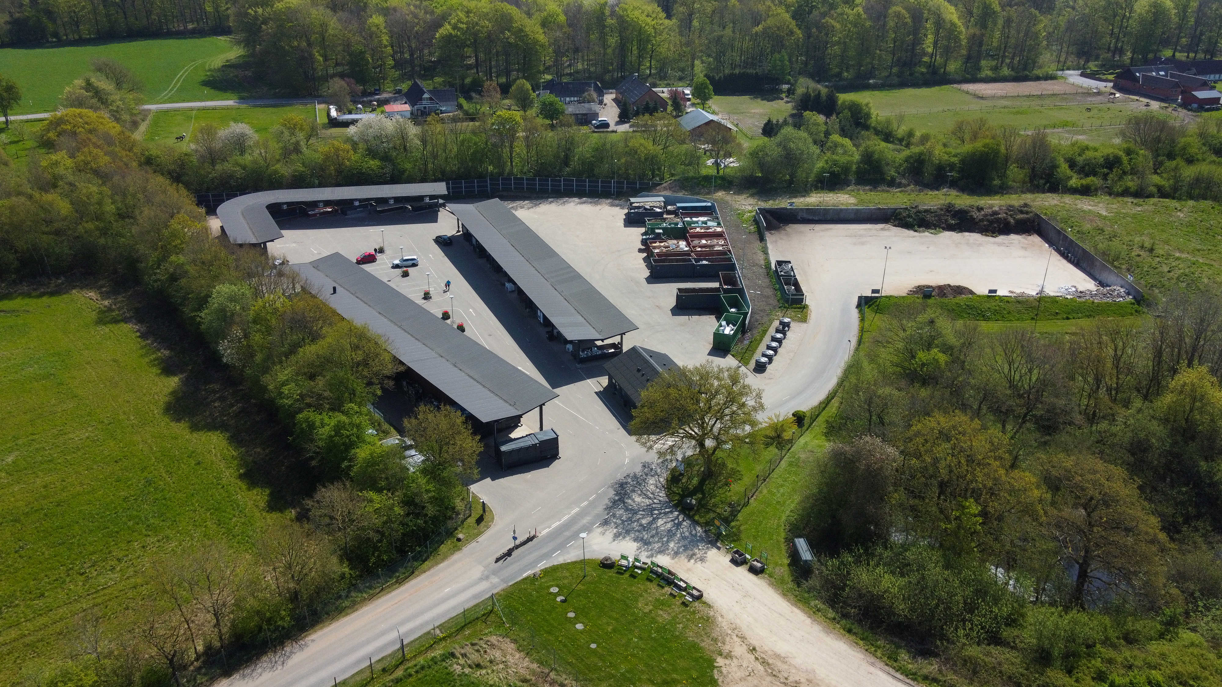 Luftfoto af Børkop Genbrugsplads omgivet af grønne træer og landlige omgivelser. Pladsen er inddelt i overdækkede sorteringsområder og containere til forskellige affaldstyper.