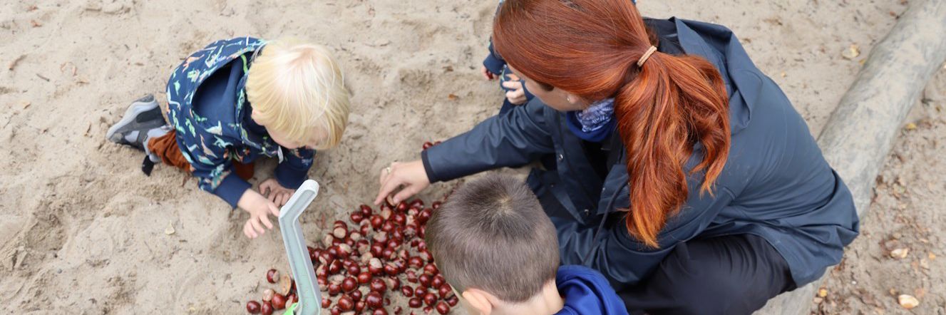 Galaksen, to børn og en voksen leger med kastanjer i sand