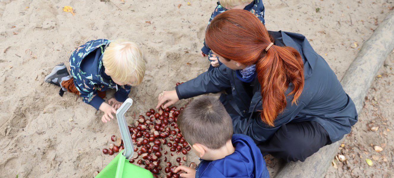 Galaksen, to børn og en voksen leger med kastanjer i sand