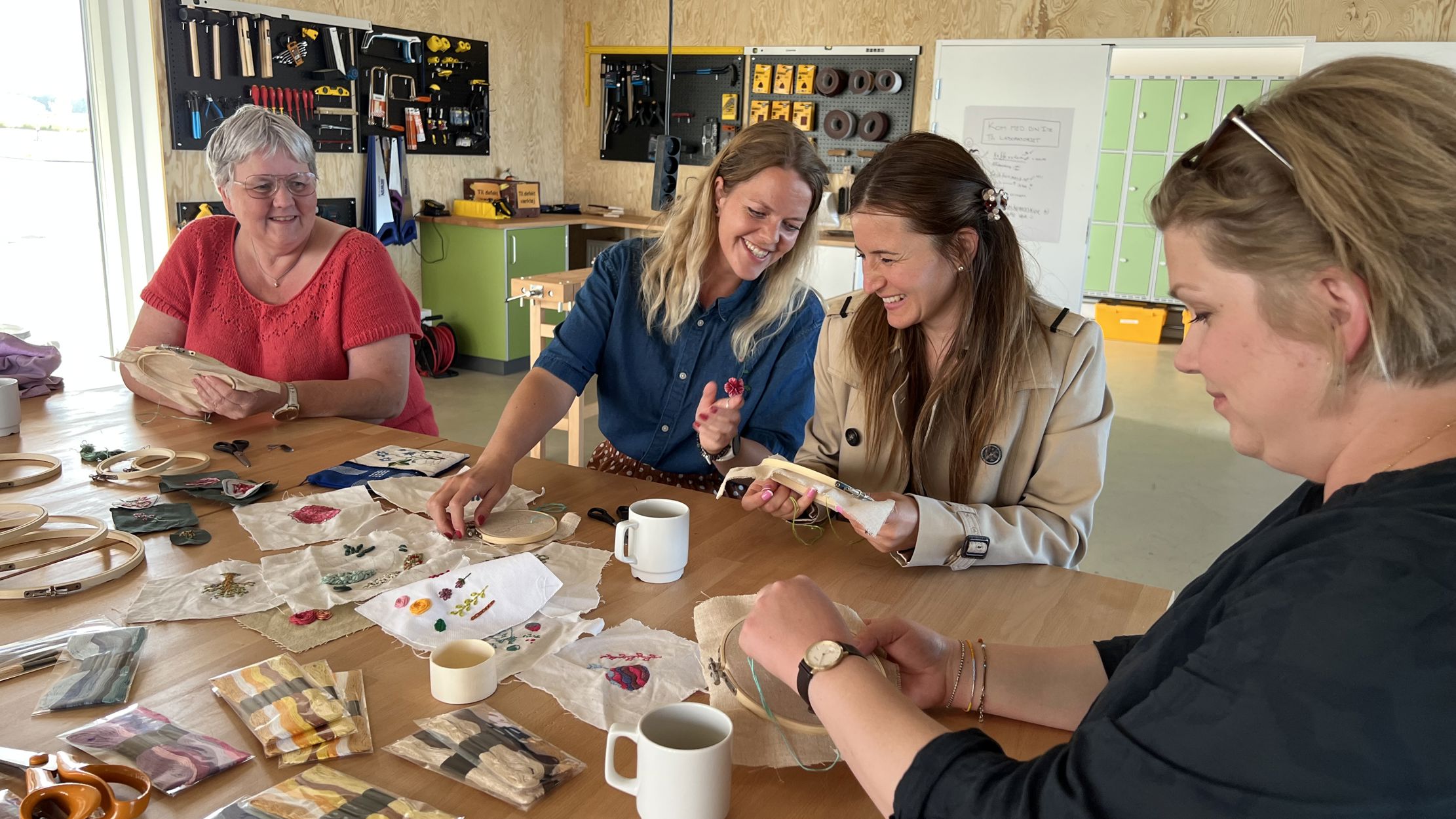 Fire smilede kvinder i alle aldre sidder ved et arbejdsbord i Laboratoriet på RCV og broderer.