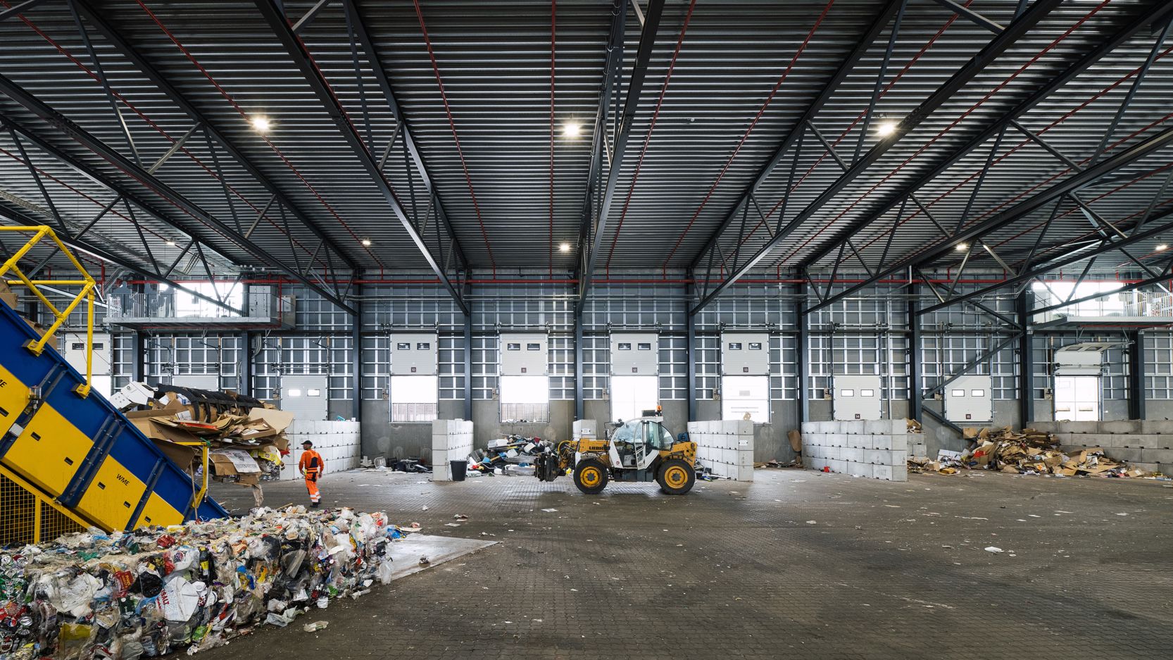 Indendørs foto af RessourceCenter Vejle's affaldssorteringshal med en gaffeltruck i arbejde og store bunker af sorteret affald, klar til videre forarbejdnin