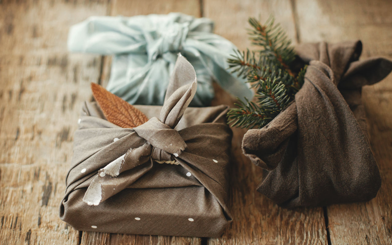 Tre julegaver pakket ind i stof med grene og blade – furoshiki-inspireret, bæredygtig indpakning.