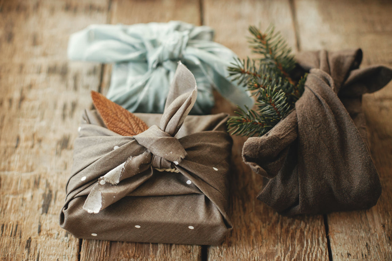 Tre julegaver pakket ind i stof med grene og blade – furoshiki-inspireret, bæredygtig indpakning.