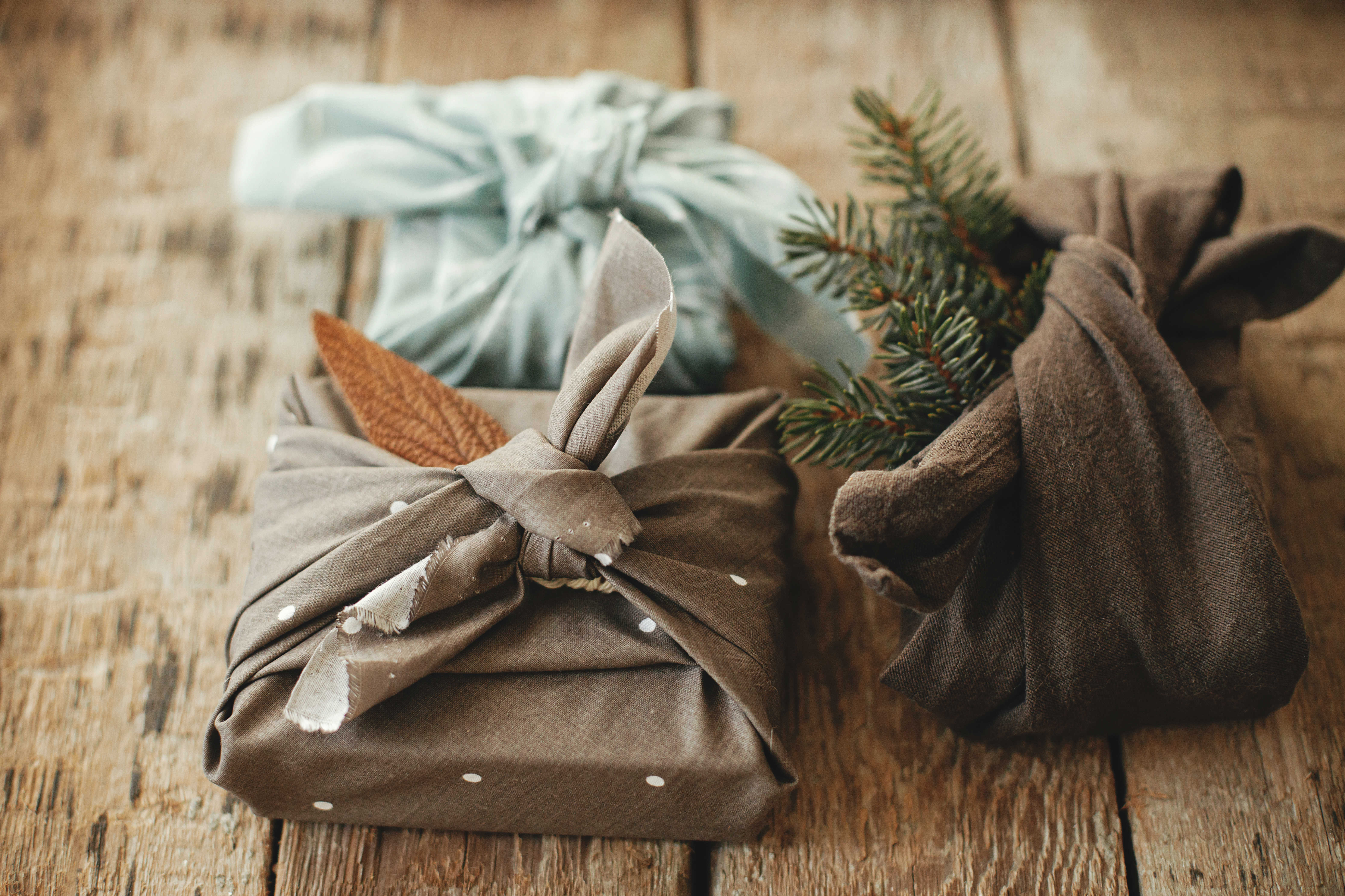 Tre julegaver pakket ind i stof med grene og blade – furoshiki-inspireret, bæredygtig indpakning.