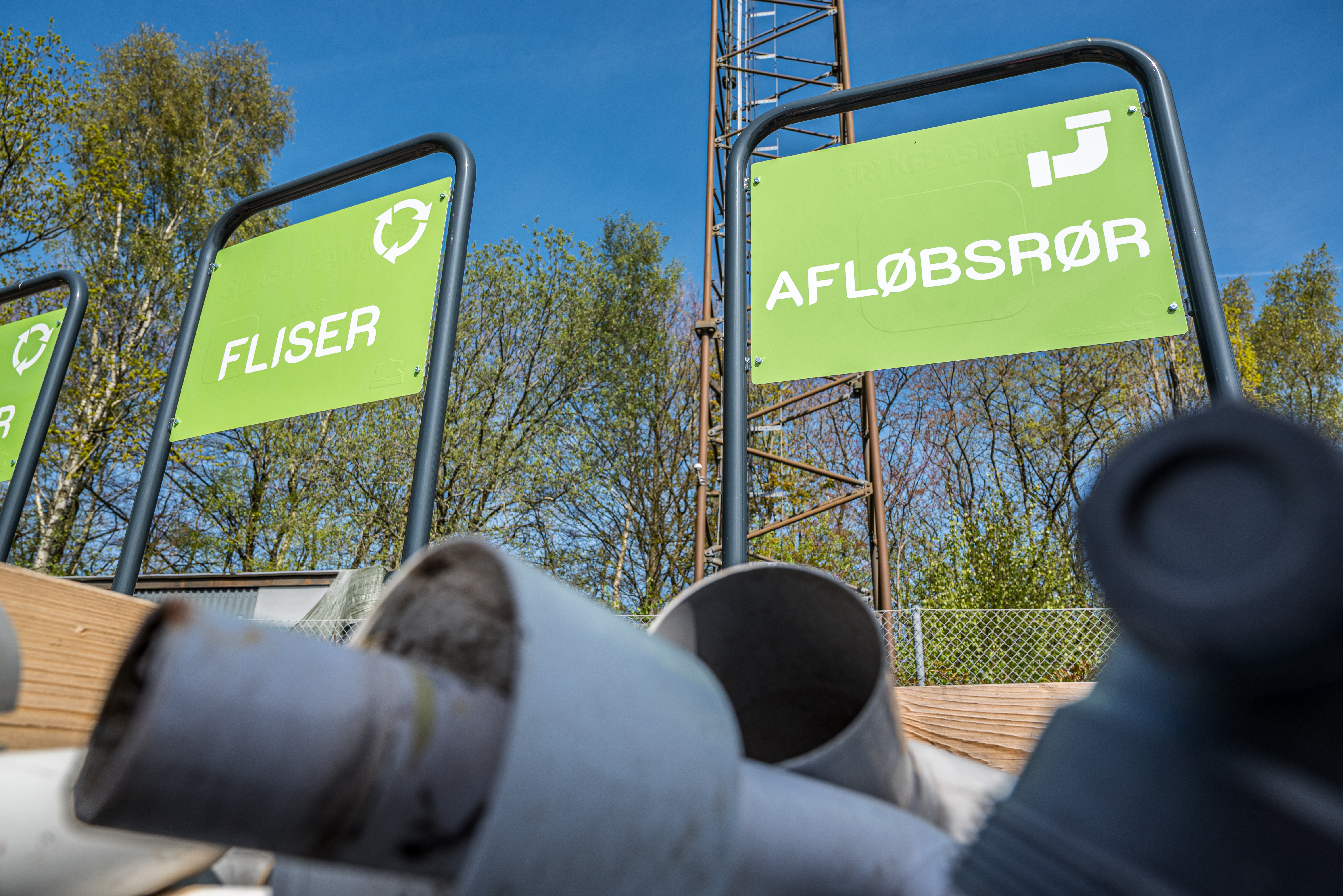 Grønt skilt med teksten 'FLISER' og 'AFLØBSRØR' over en palle med overskudsmaterialer på BygIgen-området ved Egtved Genbrugsplads.