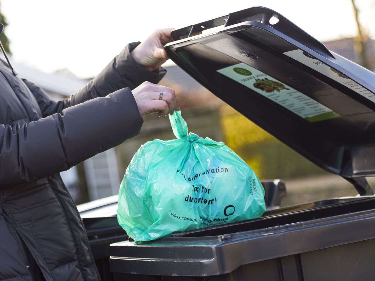 erson iført mørk jakke smider en grøn biologisk nedbrydelig pose med madaffald i en sort affaldscontainer. Containerlåget har en informationsplakat om korrekt affaldssortering. Baggrunden viser en villavej med hække og huse i sollys.