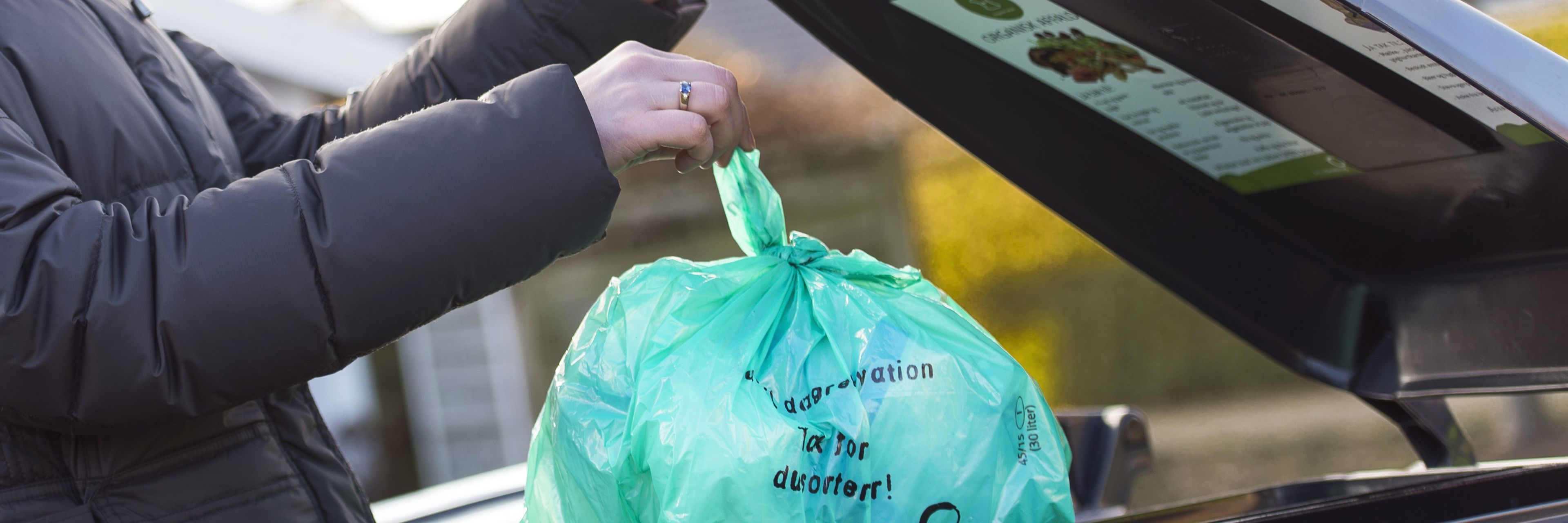 erson iført mørk jakke smider en grøn biologisk nedbrydelig pose med madaffald i en sort affaldscontainer. Containerlåget har en informationsplakat om korrekt affaldssortering. Baggrunden viser en villavej med hække og huse i sollys.