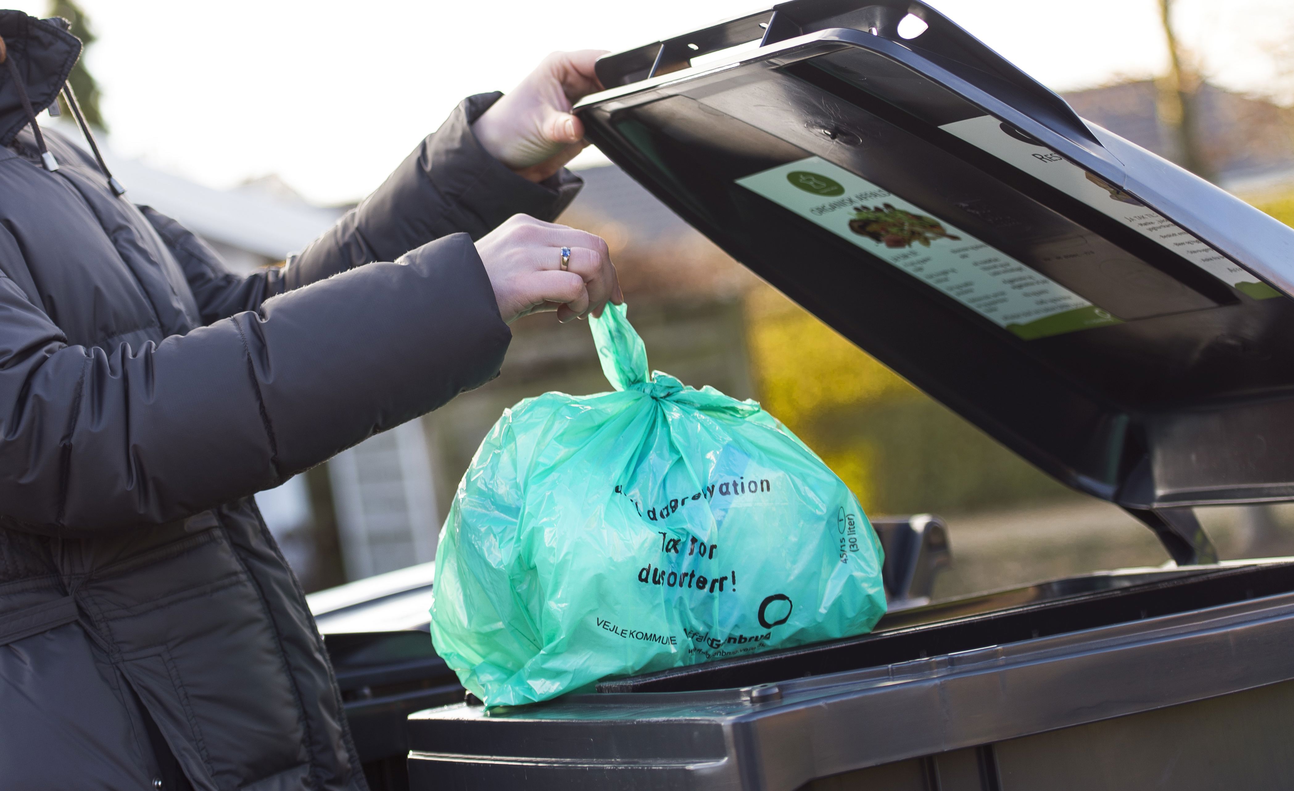 erson iført mørk jakke smider en grøn biologisk nedbrydelig pose med madaffald i en sort affaldscontainer. Containerlåget har en informationsplakat om korrekt affaldssortering. Baggrunden viser en villavej med hække og huse i sollys.