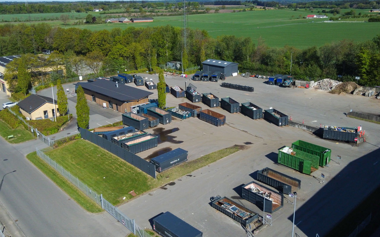 Oversigtsfoto af Egtved Genbrugsplads med udsyn til containere og sorteringsområder. Pladsen er omgivet af grønne træer og bygninger, og der er tydelige kørselsbaner til biler.