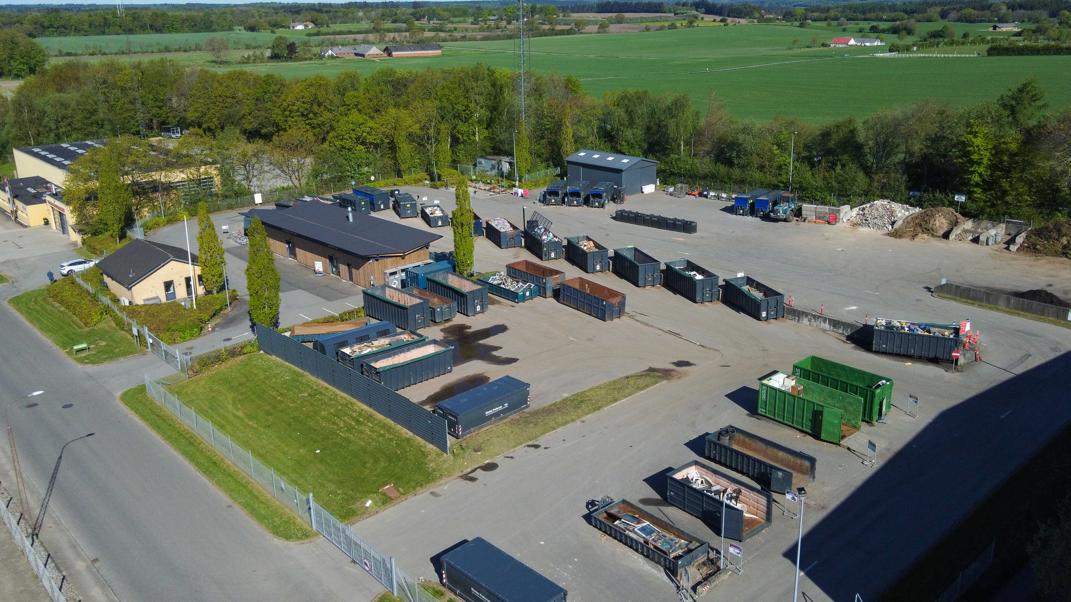 Oversigtsfoto af Egtved Genbrugsplads med udsyn til containere og sorteringsområder. Pladsen er omgivet af grønne træer og bygninger, og der er tydelige kørselsbaner til biler.