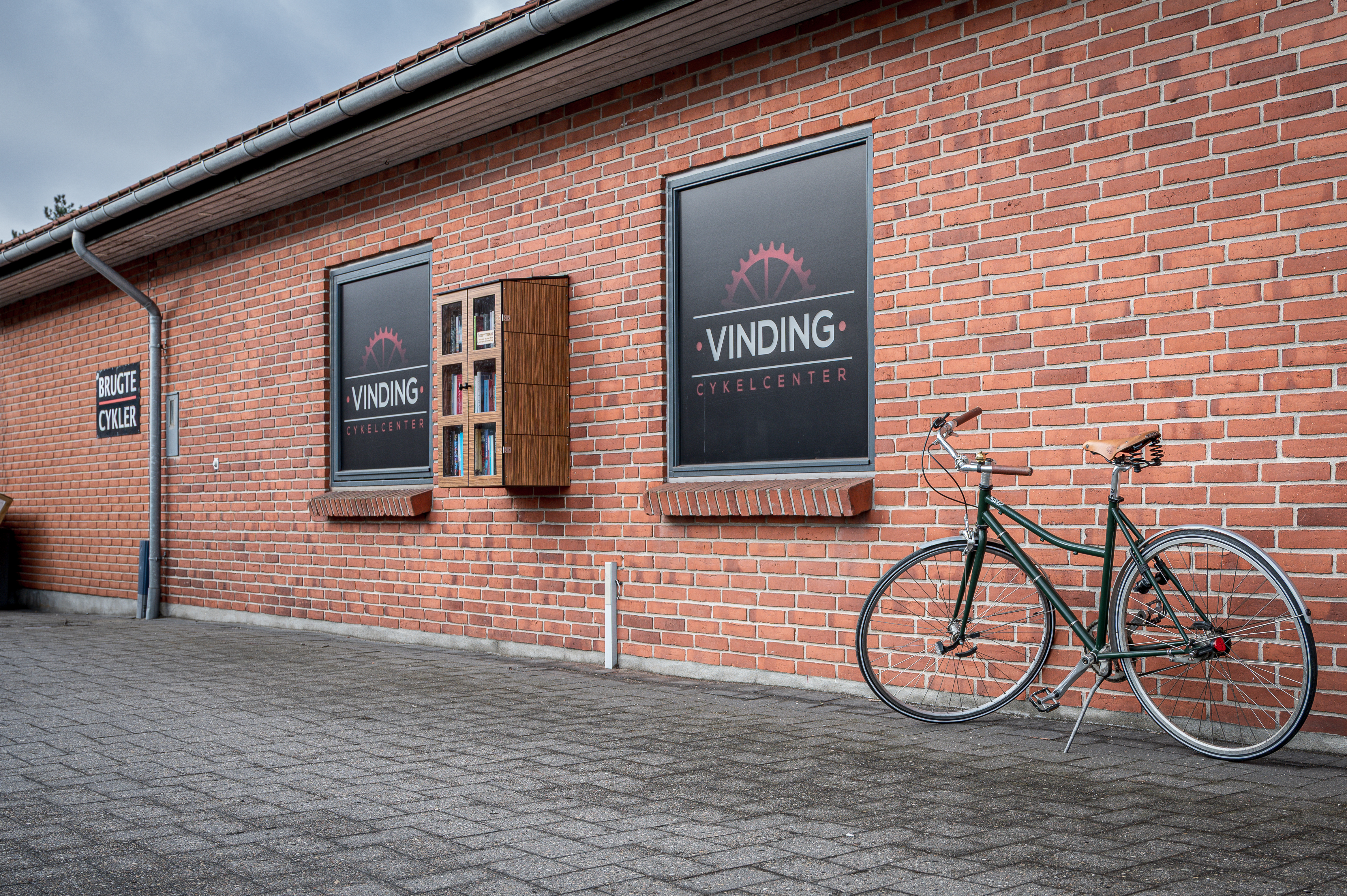 Murstensbygning med et udendørs bogskab på væggen og en cykel parkeret foran.
