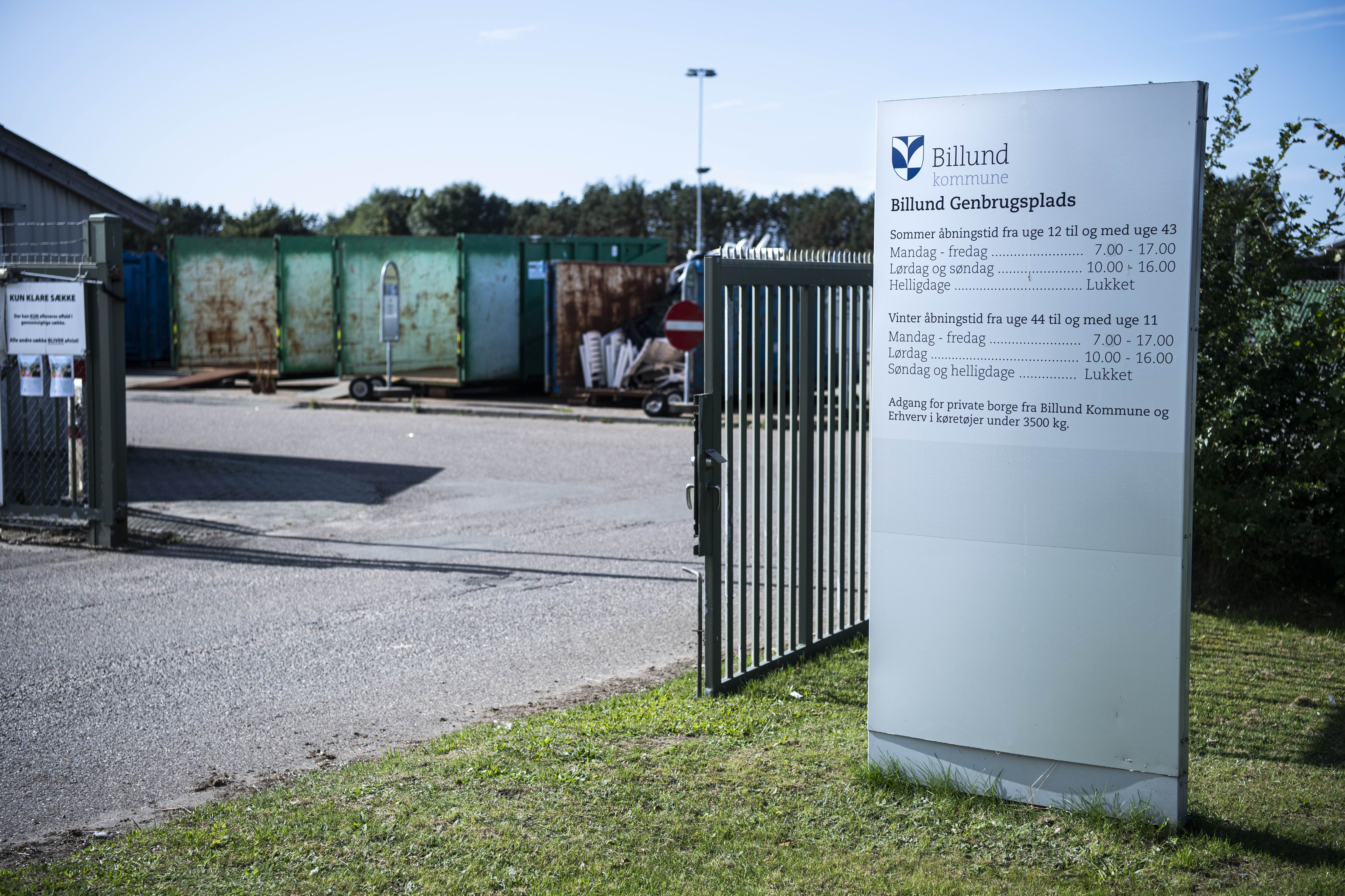 Indgang til Billund Genbrugsplads med en stor informationsstander, der viser åbningstiderne. En åben port leder ind til pladsen, hvor flere store containere til affaldssortering er synlige i baggrunden. Skilte og afspærringer markerer adgangsforholdene.