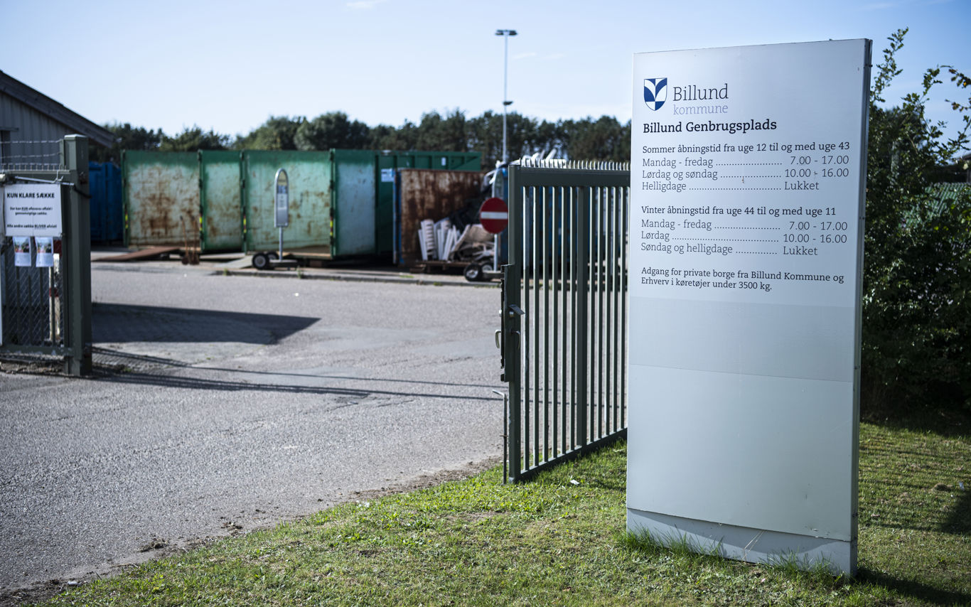 Indgang til Billund Genbrugsplads med en stor informationsstander, der viser åbningstiderne. En åben port leder ind til pladsen, hvor flere store containere til affaldssortering er synlige i baggrunden. Skilte og afspærringer markerer adgangsforholdene.