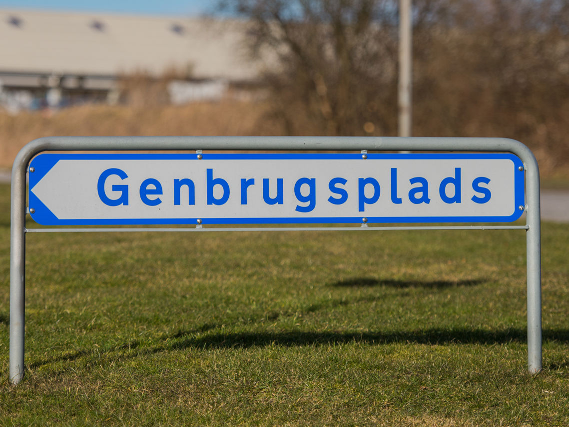 Skilt med teksten "Genbrugsplads" på en græsplæne. Skiltet er hvidt med blå kant