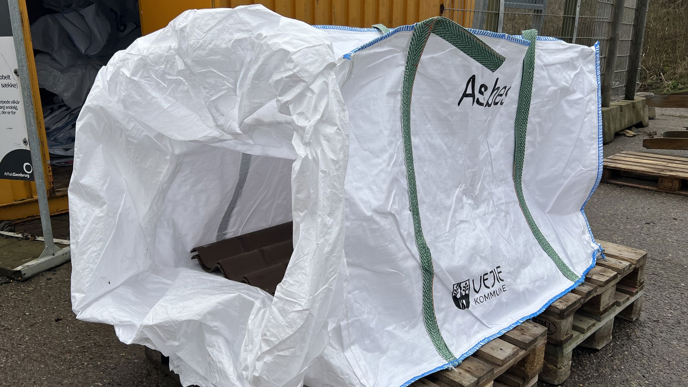 Stor hvid asbest-sæk med grønne håndtag og Vejle Kommunes logo, placeret på en træpalle ved siden af en gul container. Sækken er åben og klar til brug