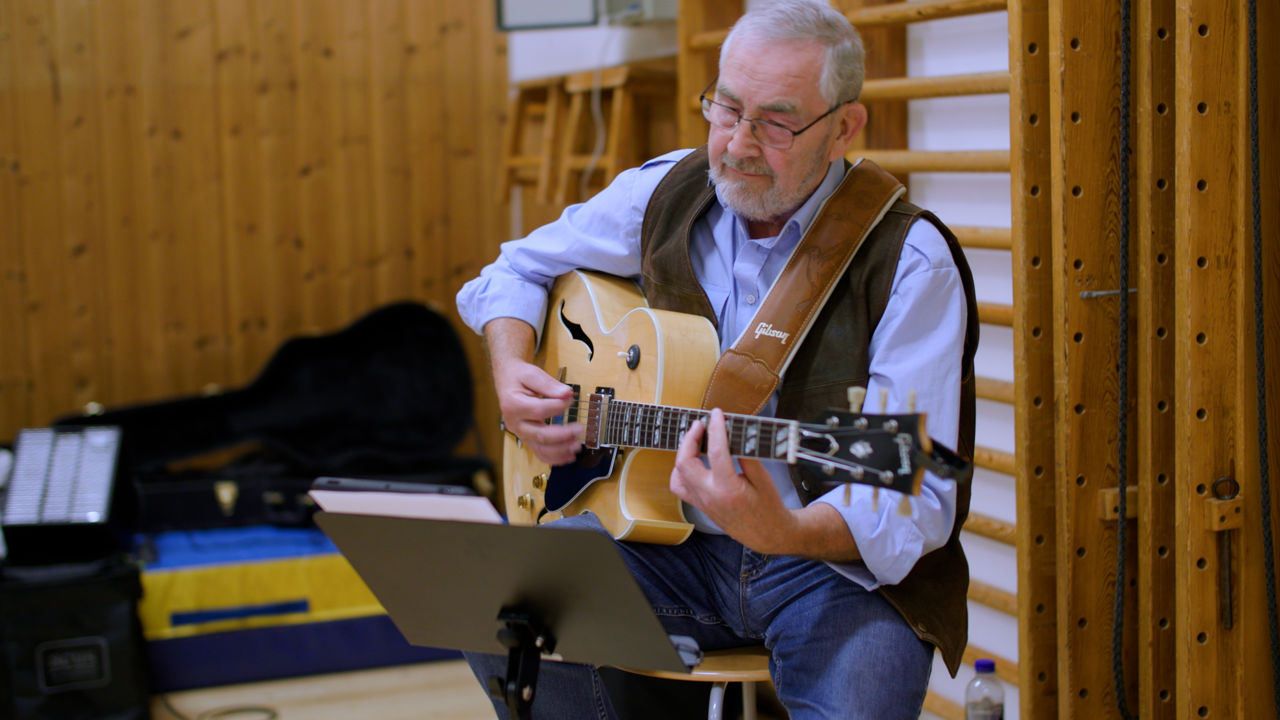 Ældre mand med guitar siddende på en skammel med nodestativ