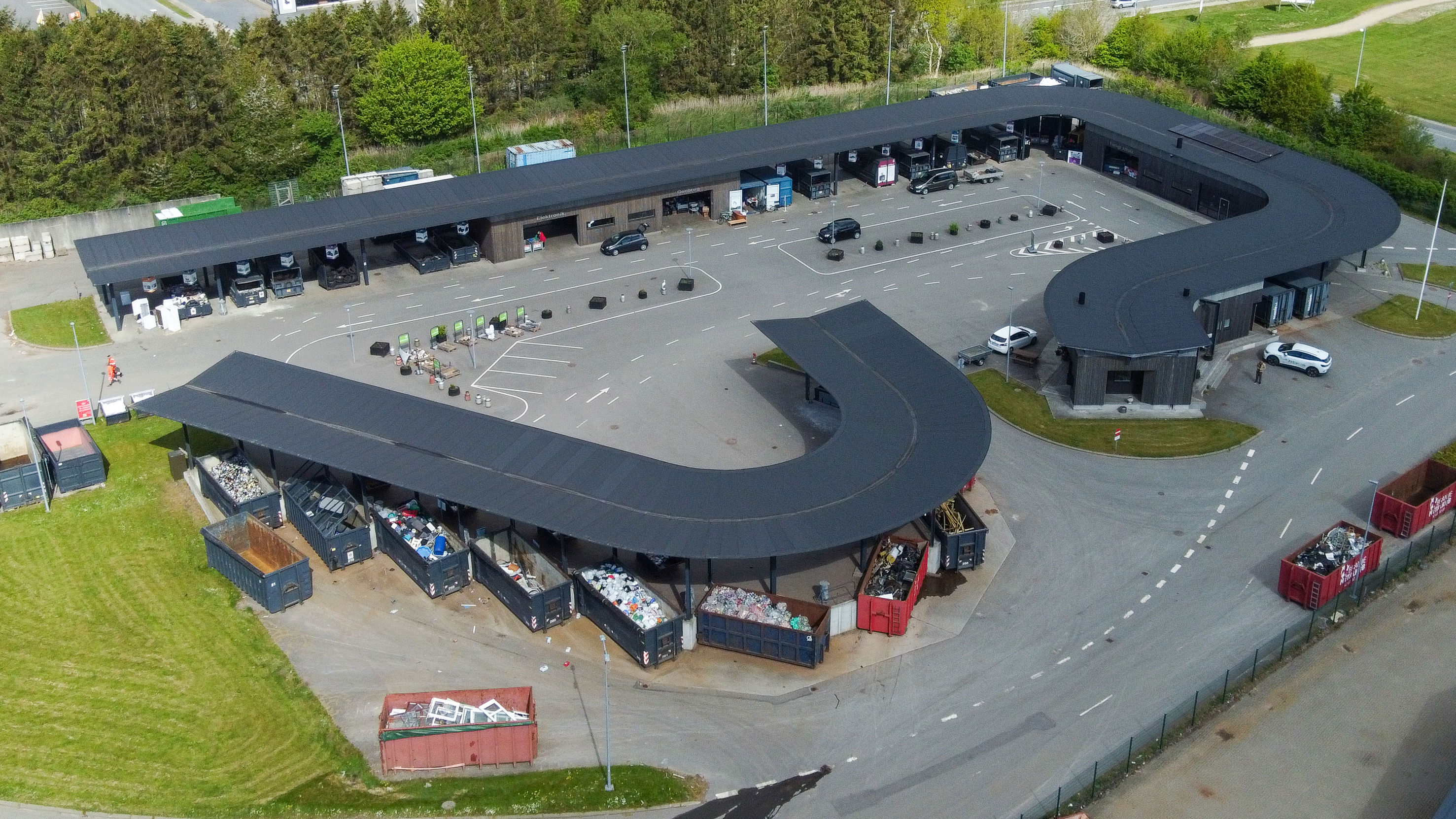 Oversigtsfoto af hele Give Genbrugsplads omgivet af grønne områder og vejnet. Billedet viser hele den buede bygning, kørselsretninger, containere og parkeringsområde.