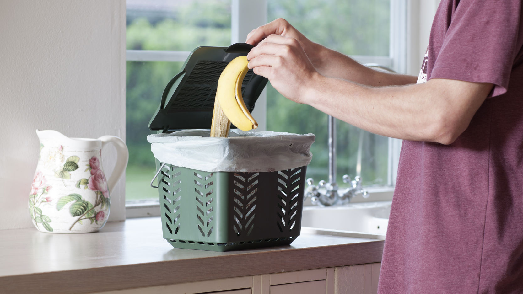 Person i t-shirt smider bananskræl i en lille grøn beholder til madaffald i et køkken med hvide skabe og træbordplade.