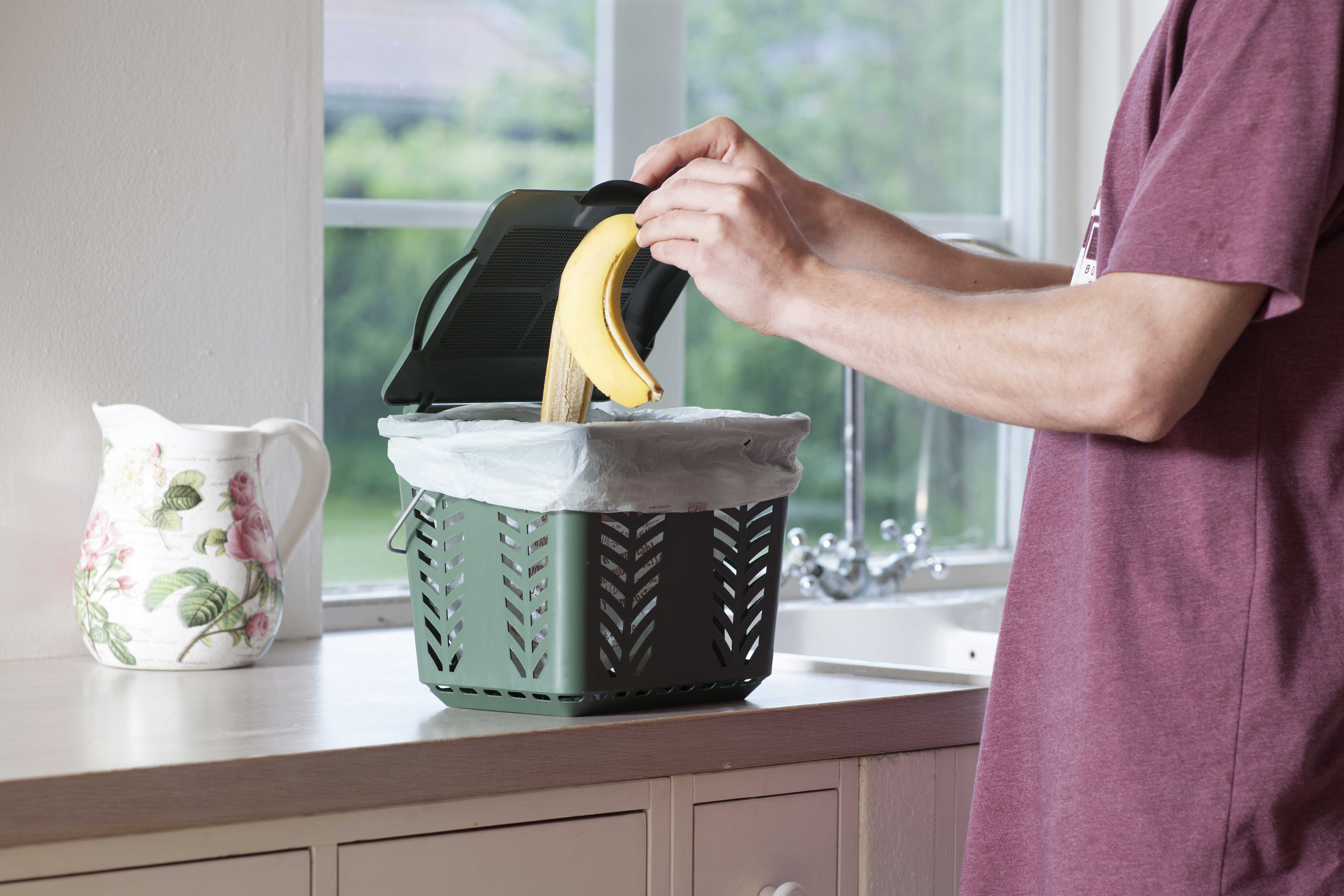 Person i t-shirt smider bananskræl i en lille grøn beholder til madaffald i et køkken med hvide skabe og træbordplade.
