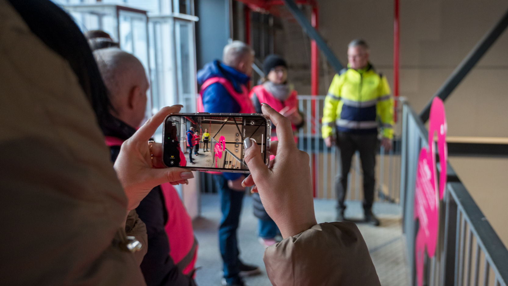 En person er i gang med at fotografere med en smartphone. På skærmen og i baggrunden ses en af platformene på Læringsbroen og flere personer i ført pink sikkerhedsveste står og taler sammen med en person i gul sikkerhedsjakke.