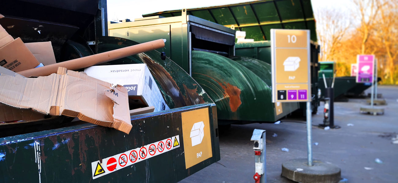 Genbrugsstation med store grønne containere til papaffald. En container i forgrunden er fyldt med papkasser og rør, og et skilt markerer området for papsortering. Flere containere til forskellige affaldsfraktioner ses i baggrunden.