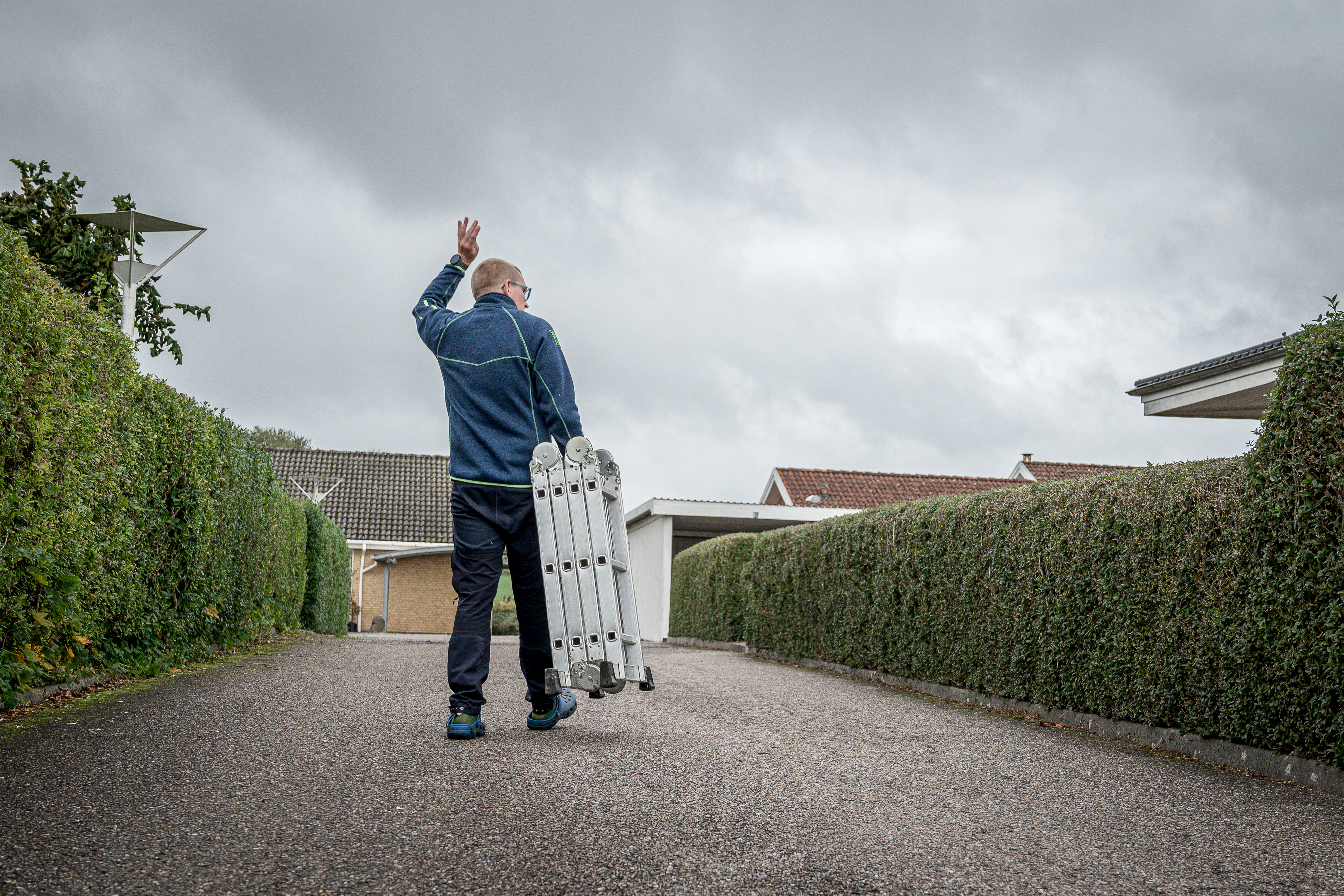 Rasmus vinker til en nabo over hækken, idet han  går ned til fællesskuret for at returnere den stige, han har lånt.