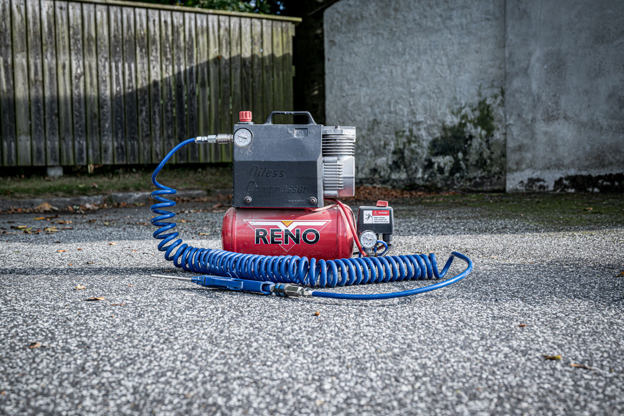 Kompressor placeret på asfalt foran et plankeværk.