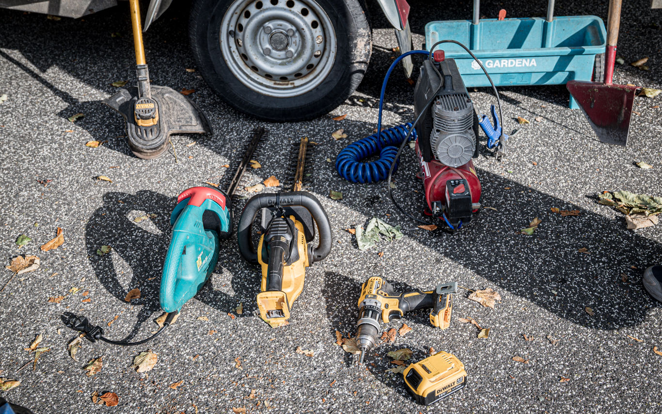 Forskelligt elværktøj og haveredskaber placeret på asfalt foran en anhænger.