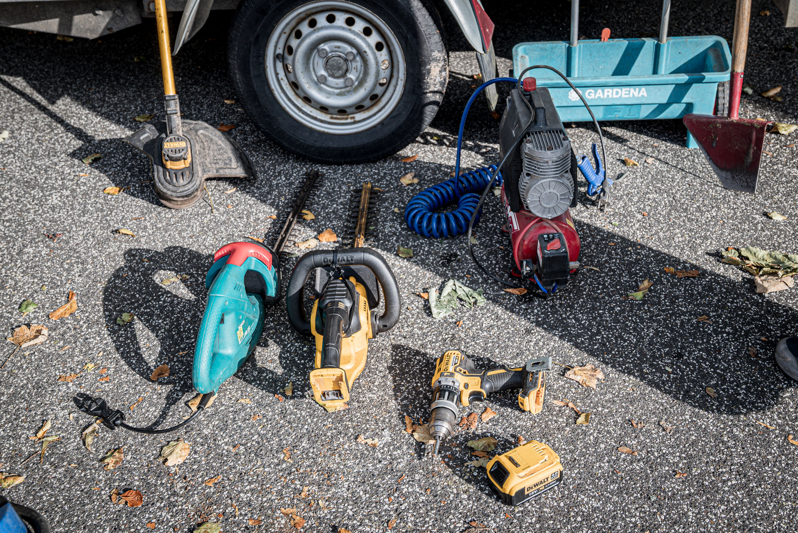 Forskelligt elværktøj og haveredskaber placeret på asfalt foran en anhænger.