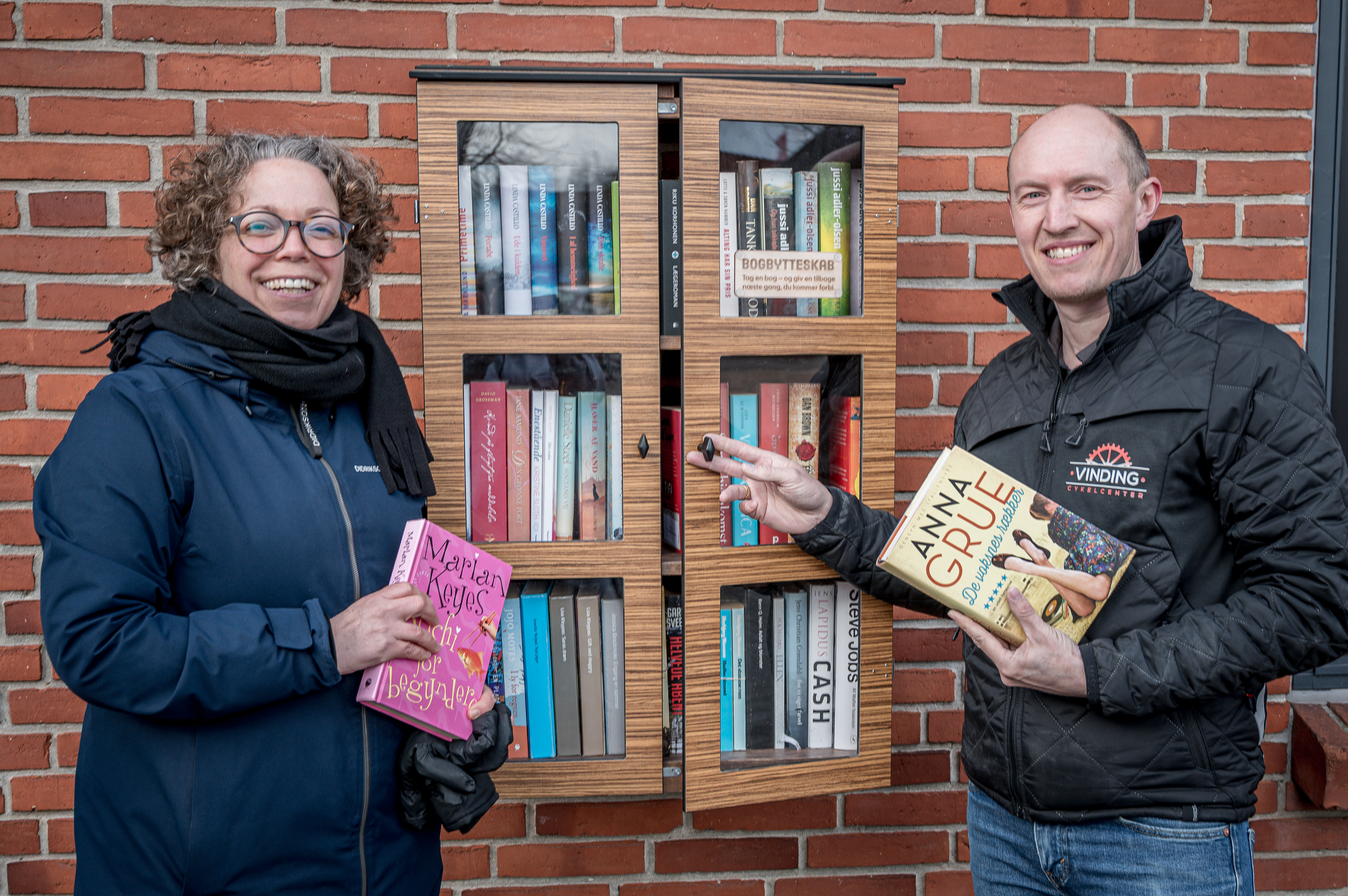 En mand og en kvinde står foran et bogskab, der hænger på en murstensvæg. De holder hver sin bog.