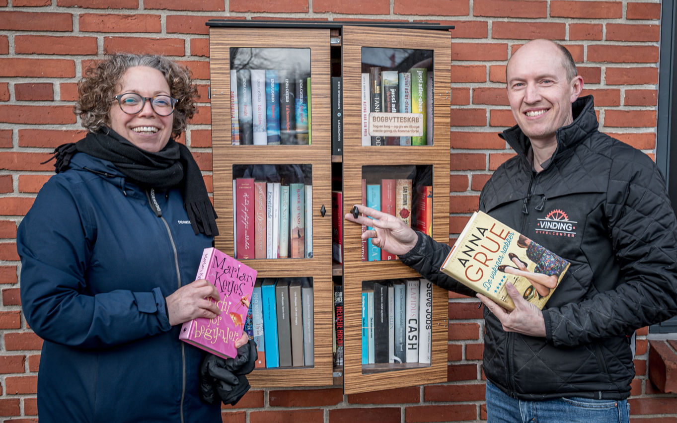 En mand og en kvinde står foran et bogskab, der hænger på en murstensvæg. De holder hver sin bog.