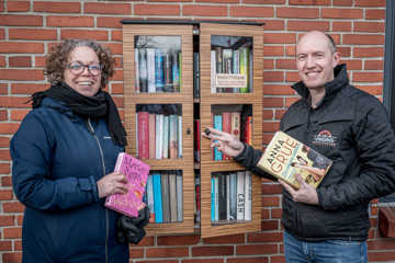 En mand og en kvinde står foran et bogskab, der hænger på en murstensvæg. De holder hver sin bog.