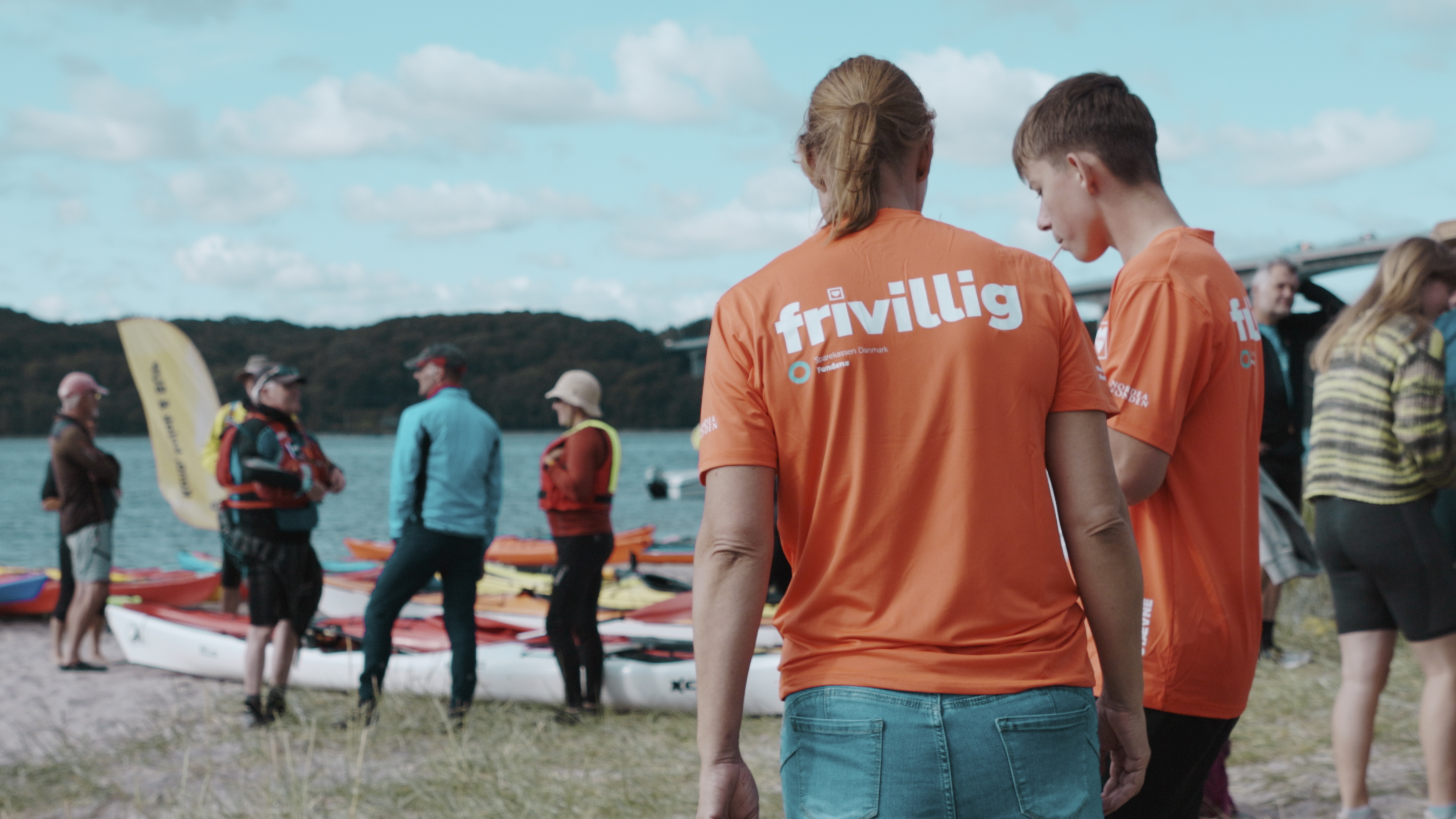 Frivillige i orange trøjer står ved Vejle Fjord med kajakker, mens en gruppe mennesker står og snakker