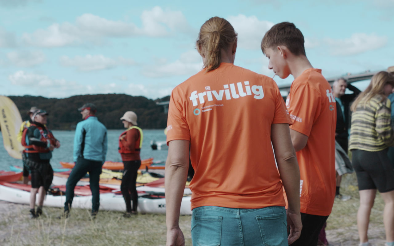 Frivillige i orange trøjer står ved Vejle Fjord med kajakker, mens en gruppe mennesker står og snakker