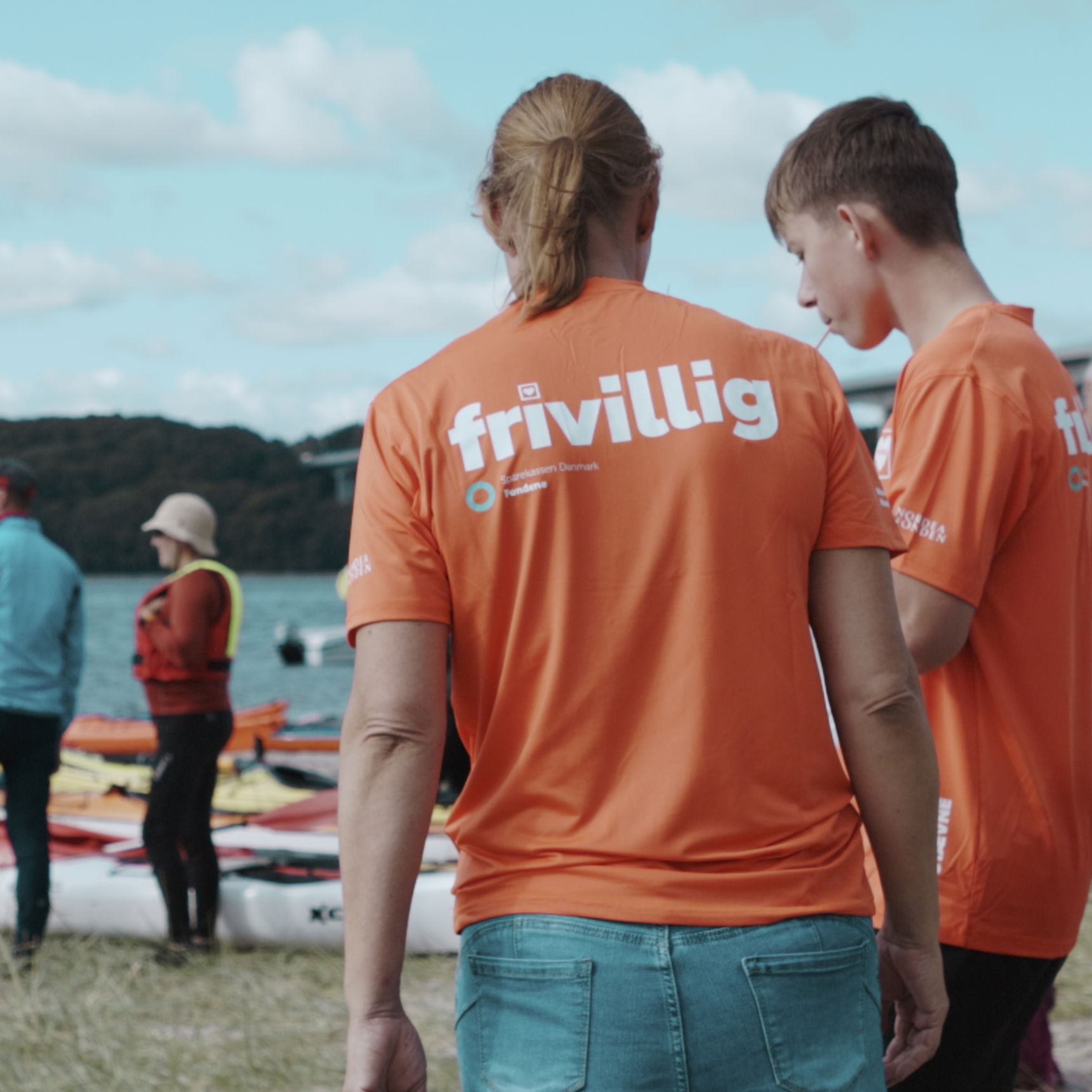 Frivillige i orange trøjer står ved Vejle Fjord med kajakker, mens en gruppe mennesker står og snakker