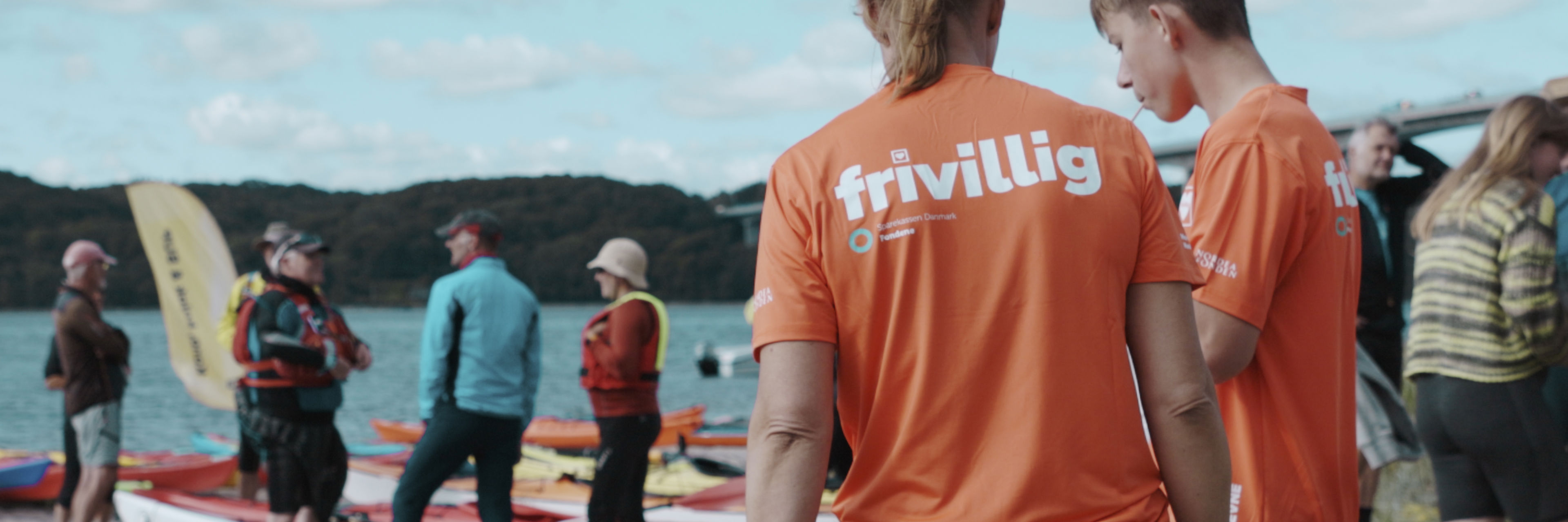 Frivillige i orange trøjer står ved Vejle Fjord med kajakker, mens en gruppe mennesker står og snakker