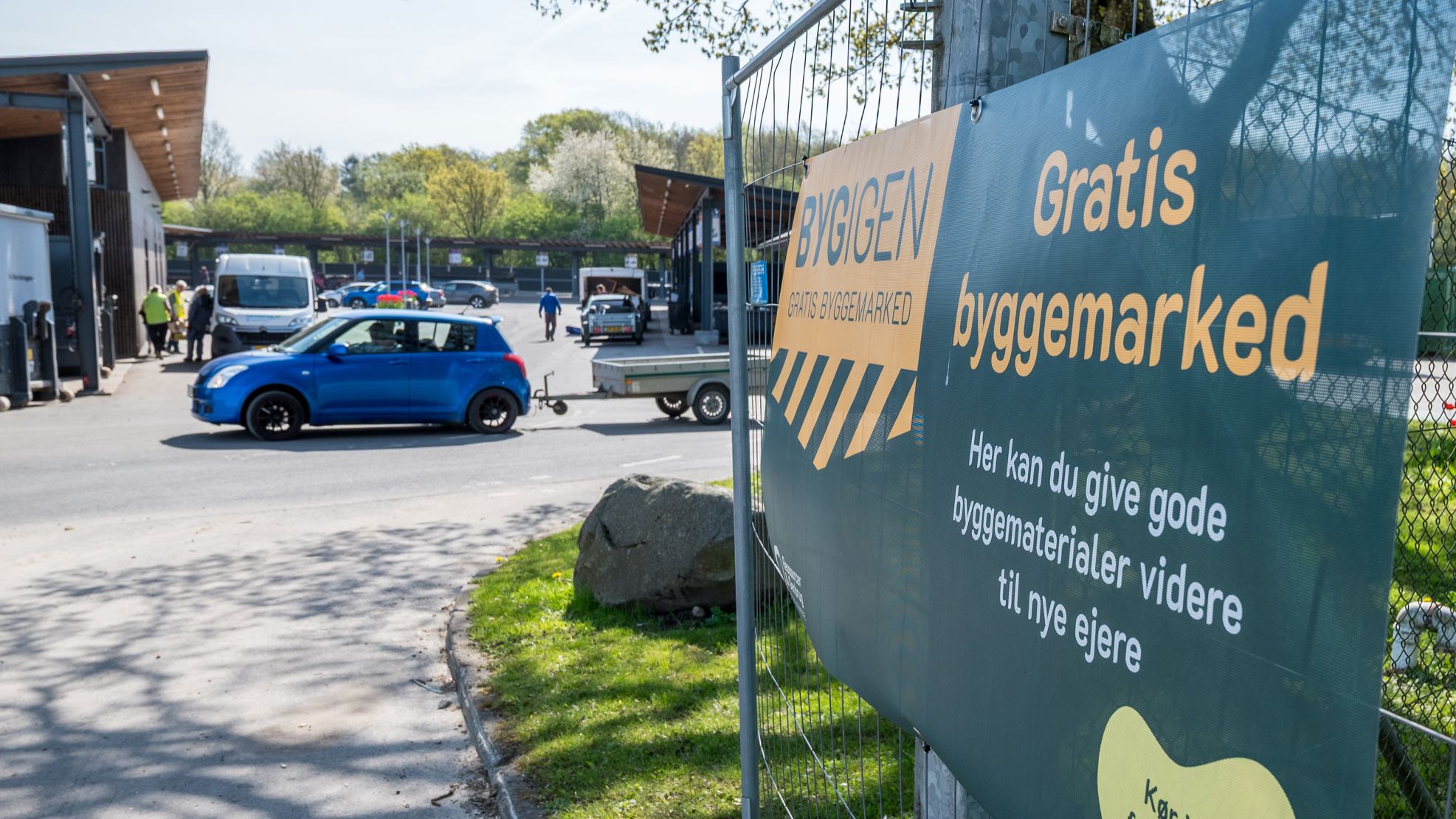 Skilt ved indgangen til BygIgen ved Børkop Genbrugsplads. Skiltet annoncerer "Gratis byggemarked" og inviterer borgere til at aflevere eller afhente gode byggematerialer, der kan genbruges.