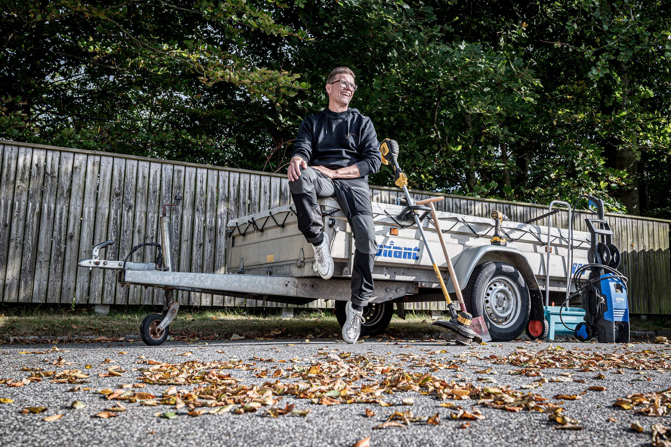 Tomas sidder på kanten af en anhænger og smiler omringet af haveredskaber og værktøj.