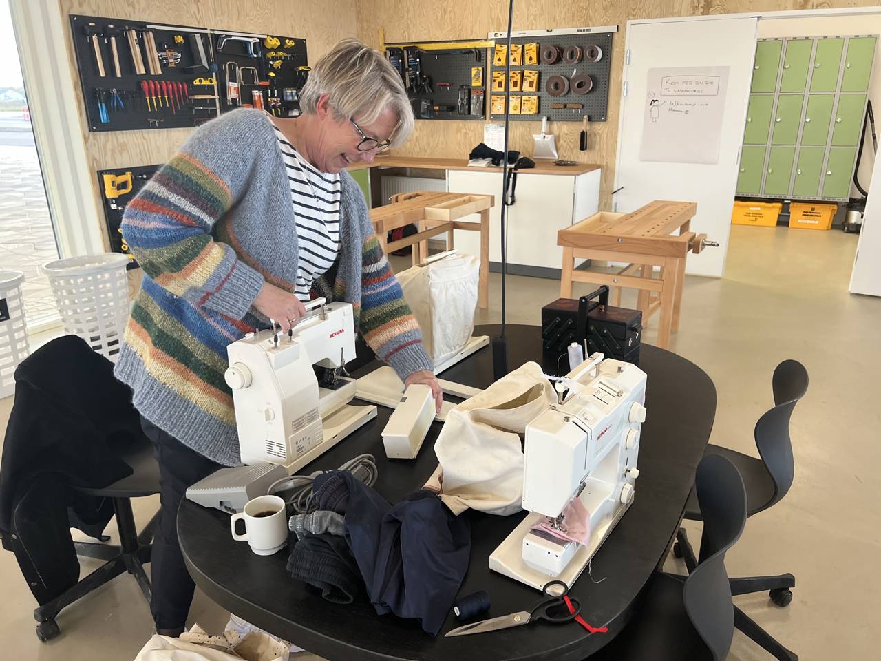 Smilende kvinde ved symaskine i Laboratoriet på RCV. I baggrunden ses træværkstedshjørnet med slagbænke og værktøj.