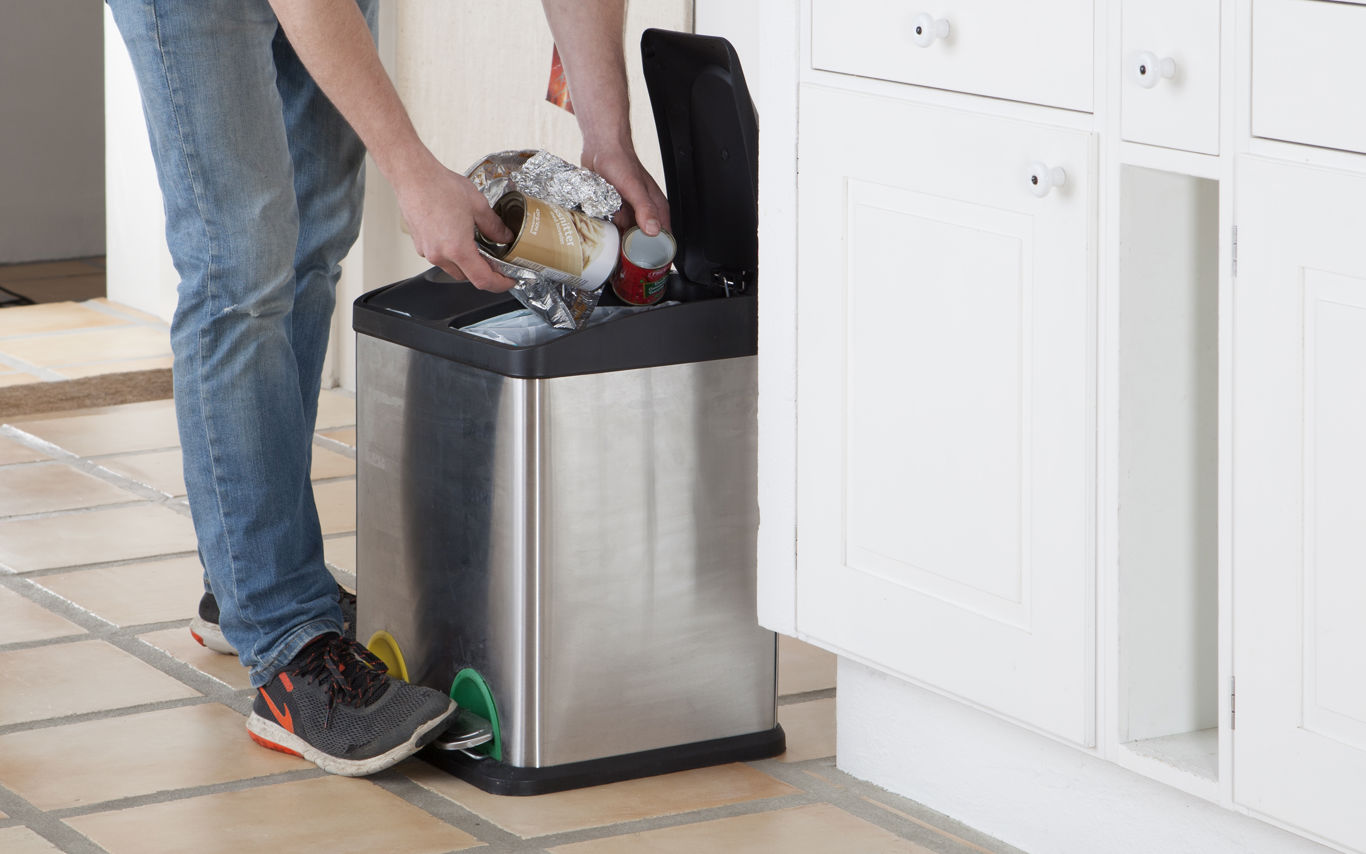 Person i jeans og sneakers smider affald i en skraldespand af rustfrit stål i et køkken med hvide skabe og flisegulv.