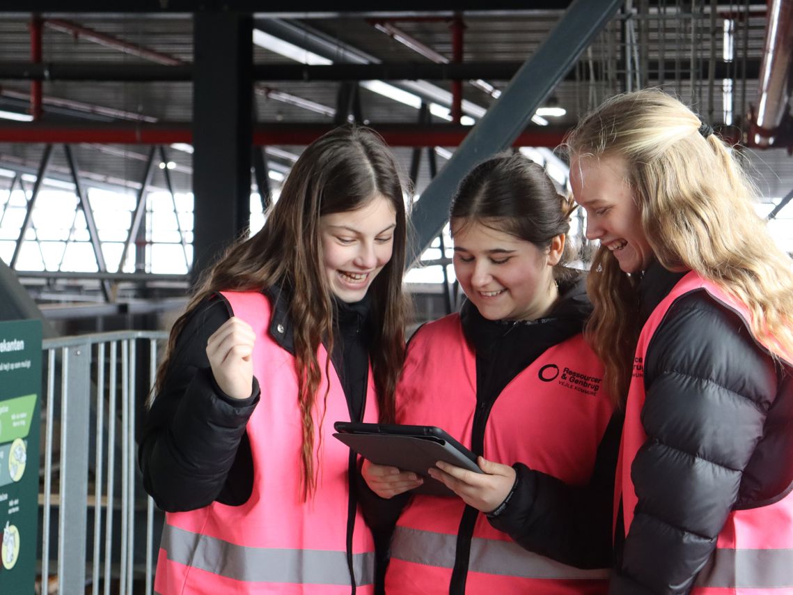 Tre piger fra 6. klasse iført pink veste kigger engageret på en iPad under et undervisningsforløb om affald og genbrug.