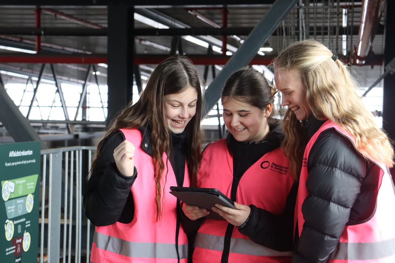 Tre piger fra 6. klasse iført pink veste kigger engageret på en iPad under et undervisningsforløb om affald og genbrug.