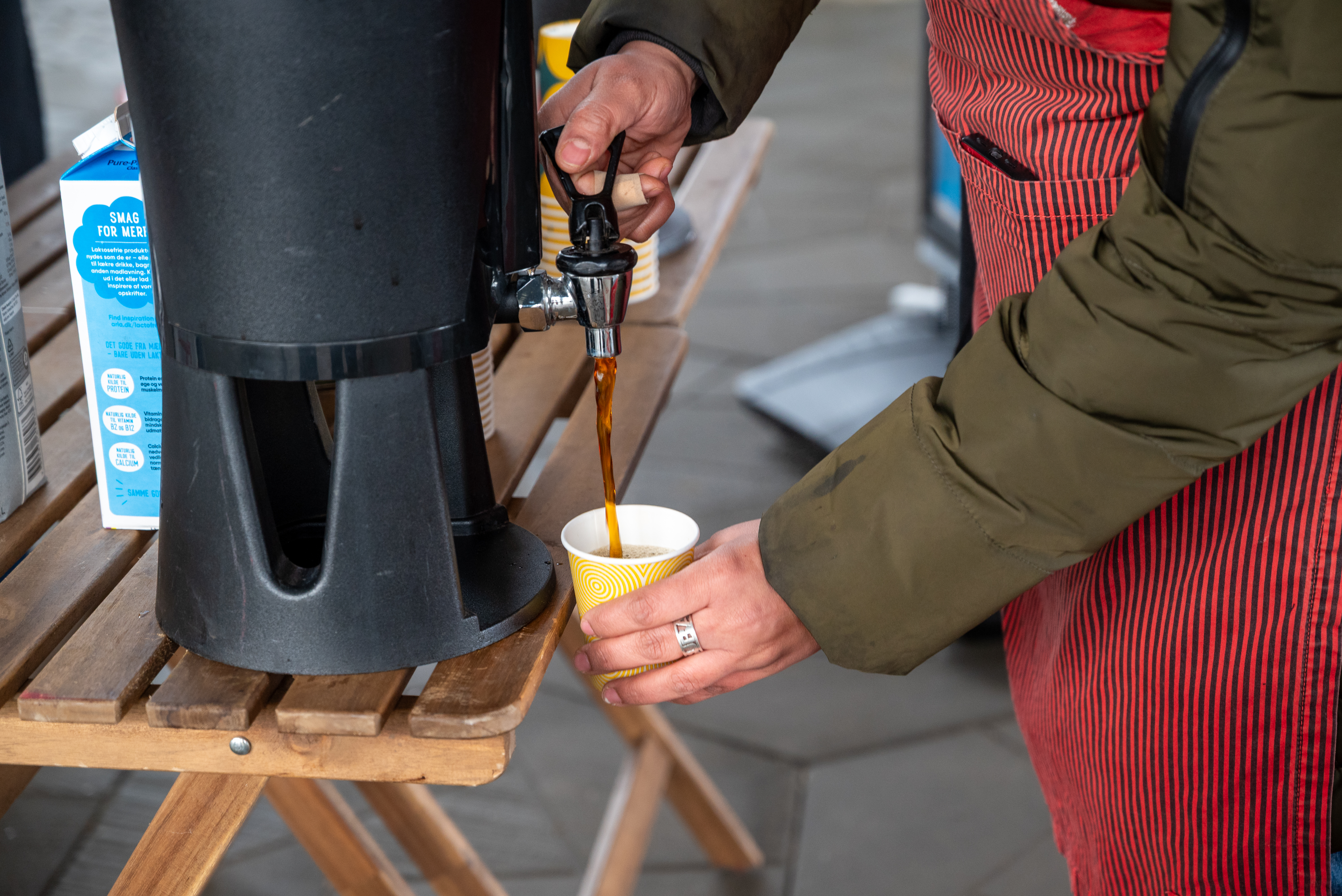 Person iført grøn jakke og rødt forklæde hælder kaffe i et krus på Torvet ved RessourceCenter Vejle i forbindelsen med fejringen af RCV's 2-års fødselsdag-