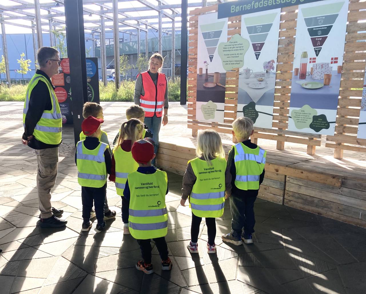 Genbrugsformidler iført pink sikkerhedsvest på Torvet ved RCV sammen med en gruppe børnehavebørn, der bliver vist rundt.