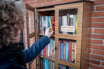 Person rækker ind i et udendørs bogskab med bøger, der er monteret på en murstensvæg.